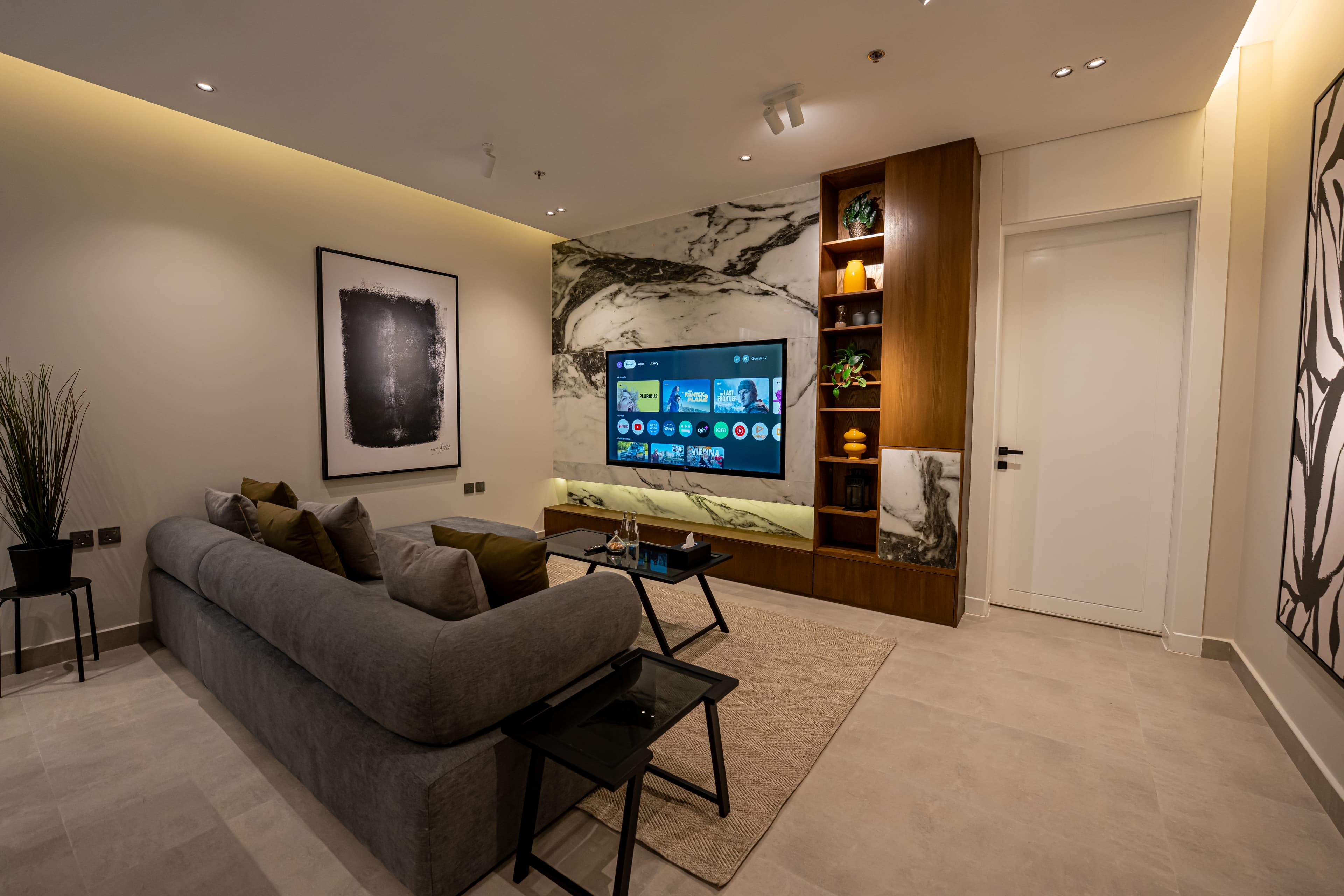 Modern living room with a grey sofa, marble TV wall, wooden shelves, and abstract wall art.