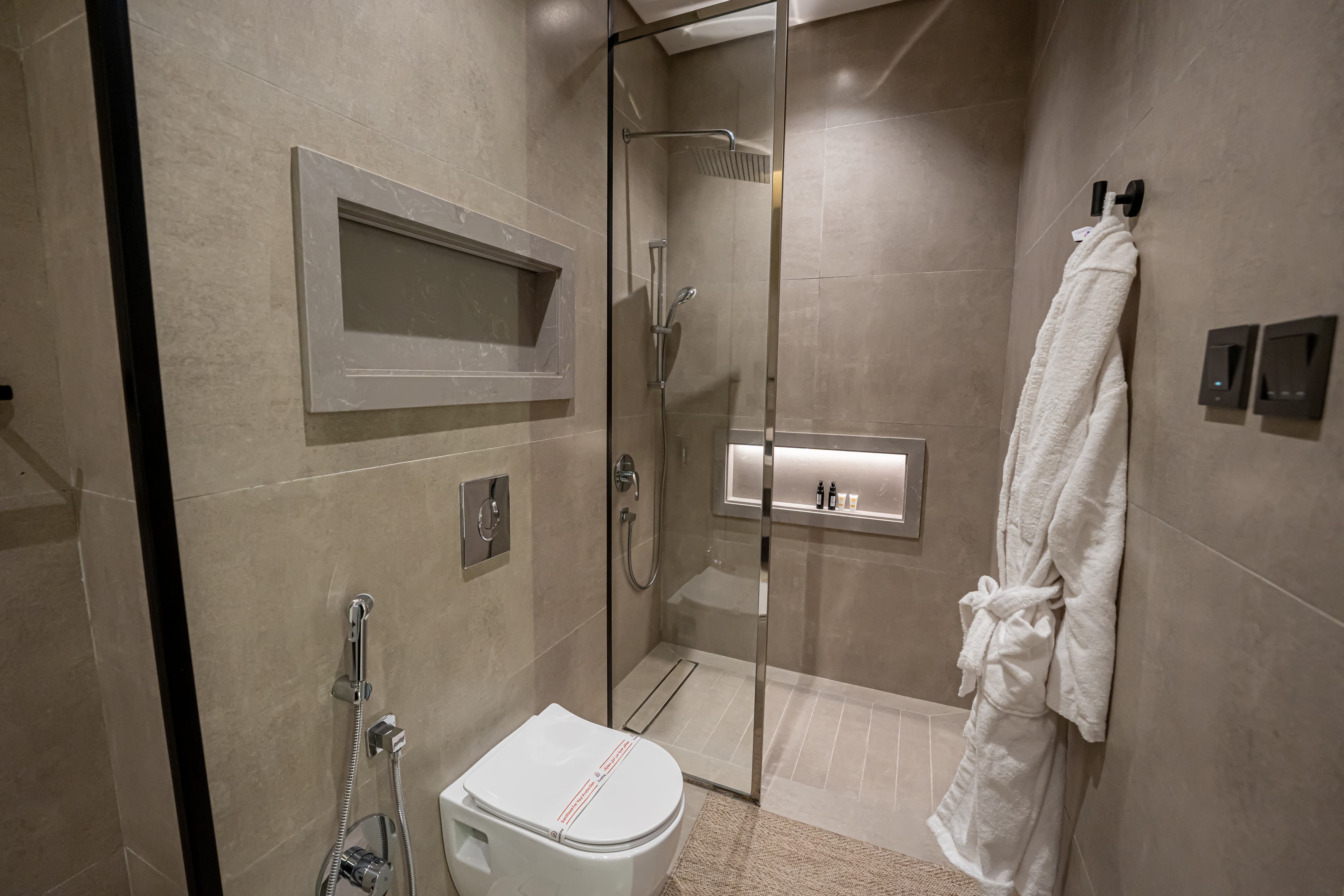 Modern beige bathroom featuring a glass shower, wall-mounted toilet, and a hanging white bathrobe.