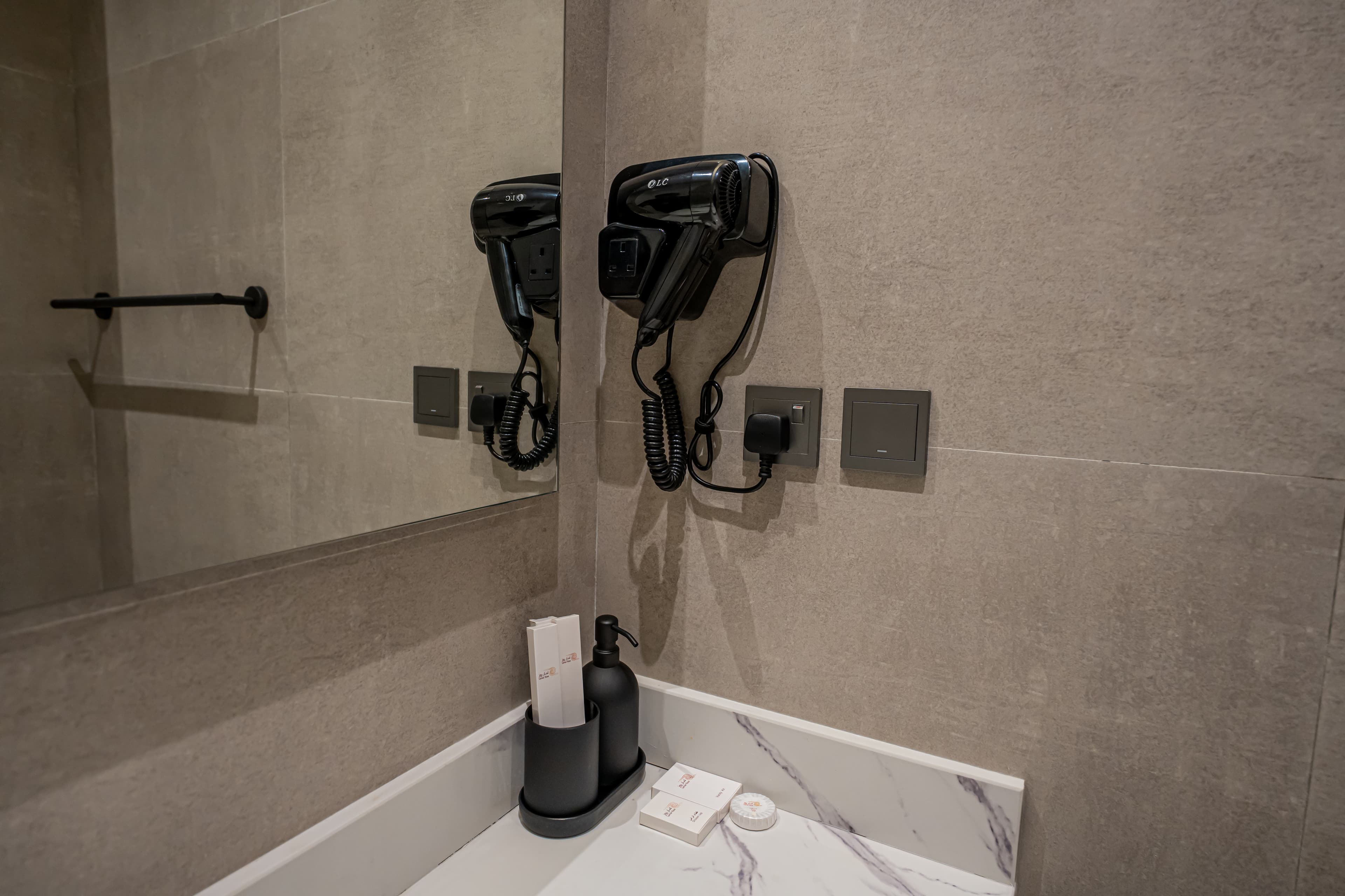 Black hair dryer mounted on a grey tiled wall above a marble vanity with toiletries.