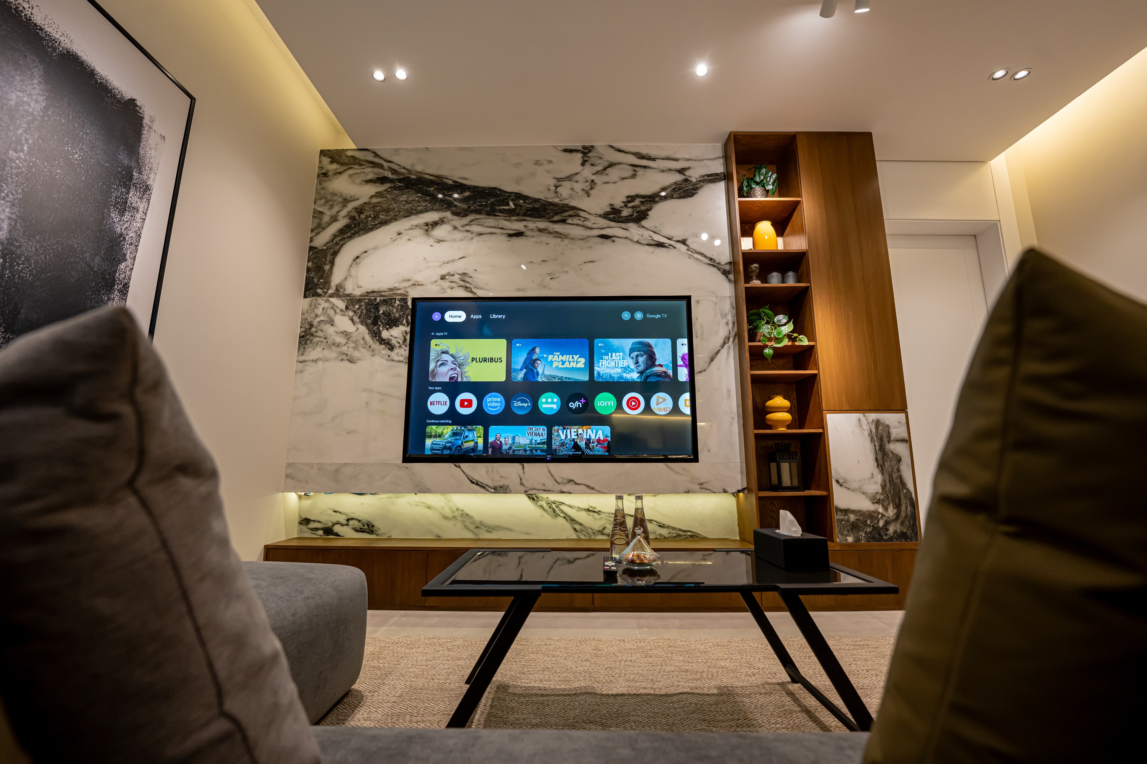 Modern living room with a TV on a marble wall, wooden shelving, and coffee table.