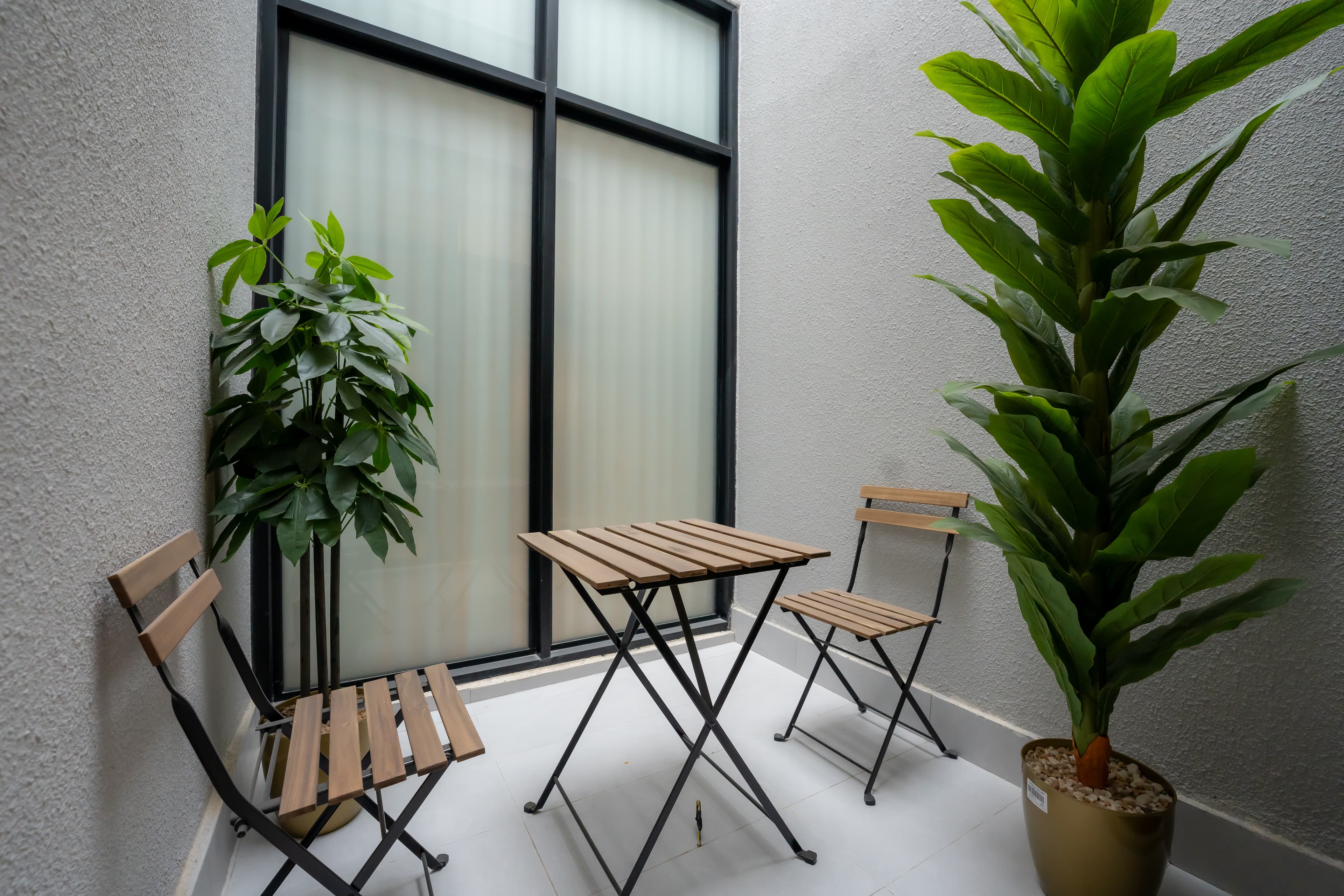 Small patio featuring a wooden bistro set, two large green plants, and a frosted window.